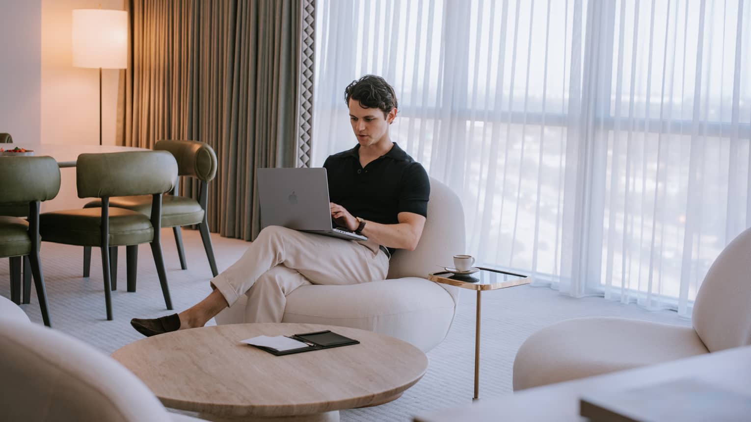 Person sitting in a living room with floor-to-ceiling windows and typing on a laptop.