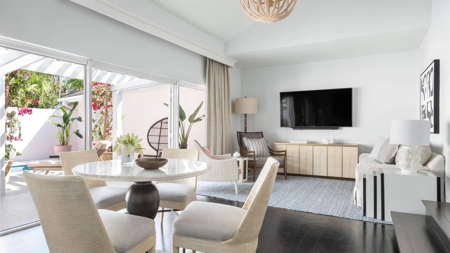 Bright resort living area with a dining table, sofa, wall-mounted TV, and sliding glass doors leading to an outdoor patio
