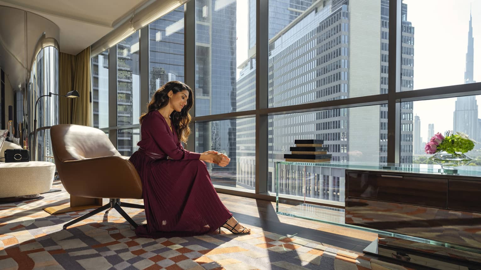 Woman in long burgundy dress sits next to floor-to-ceiling windows in sunny suite