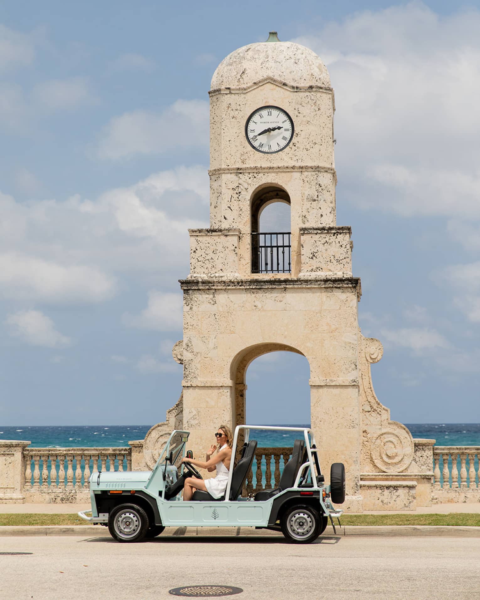 A person drives a light blue open-top vehicle past a historic clock tower by the sea. The backdrop features clear skies and the ocean.