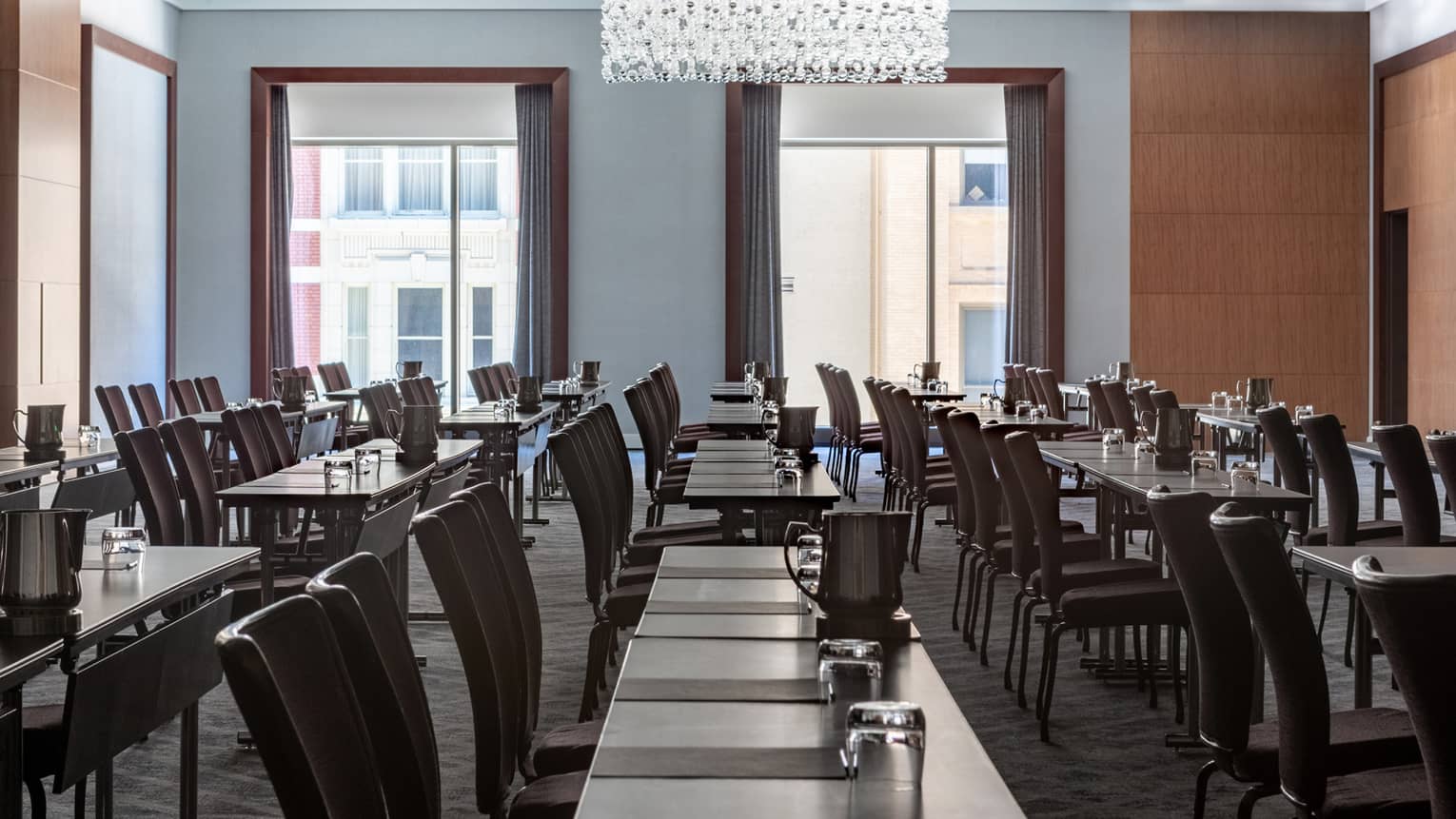 A meeting room with rows of tables and black chairs, there is also a large chandelier and large windows.
