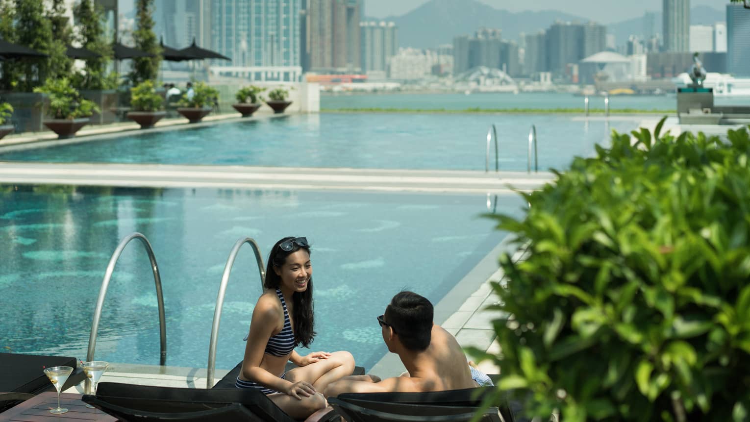 Two people are by the pool edge, and the city skyline is behind them. The pool area has greenery, lounge chairs and plants and there is a clear sky overhead.