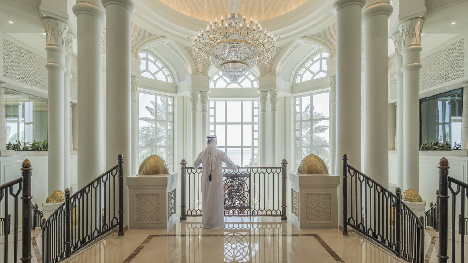 A man in a white thobe in front of enormous floor-to-ceiling windows in a marble foyer.