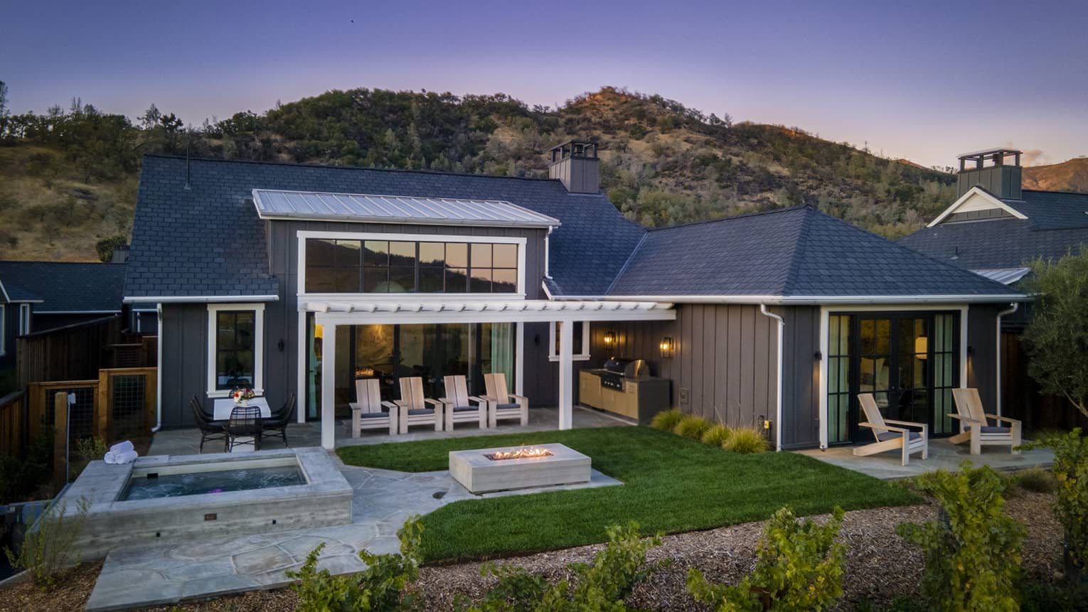 Luxury resort villa with outdoor hot tub, fire pit, lounge chairs, covered patio and grill area, surrounded by greenery and hillside views at sunset