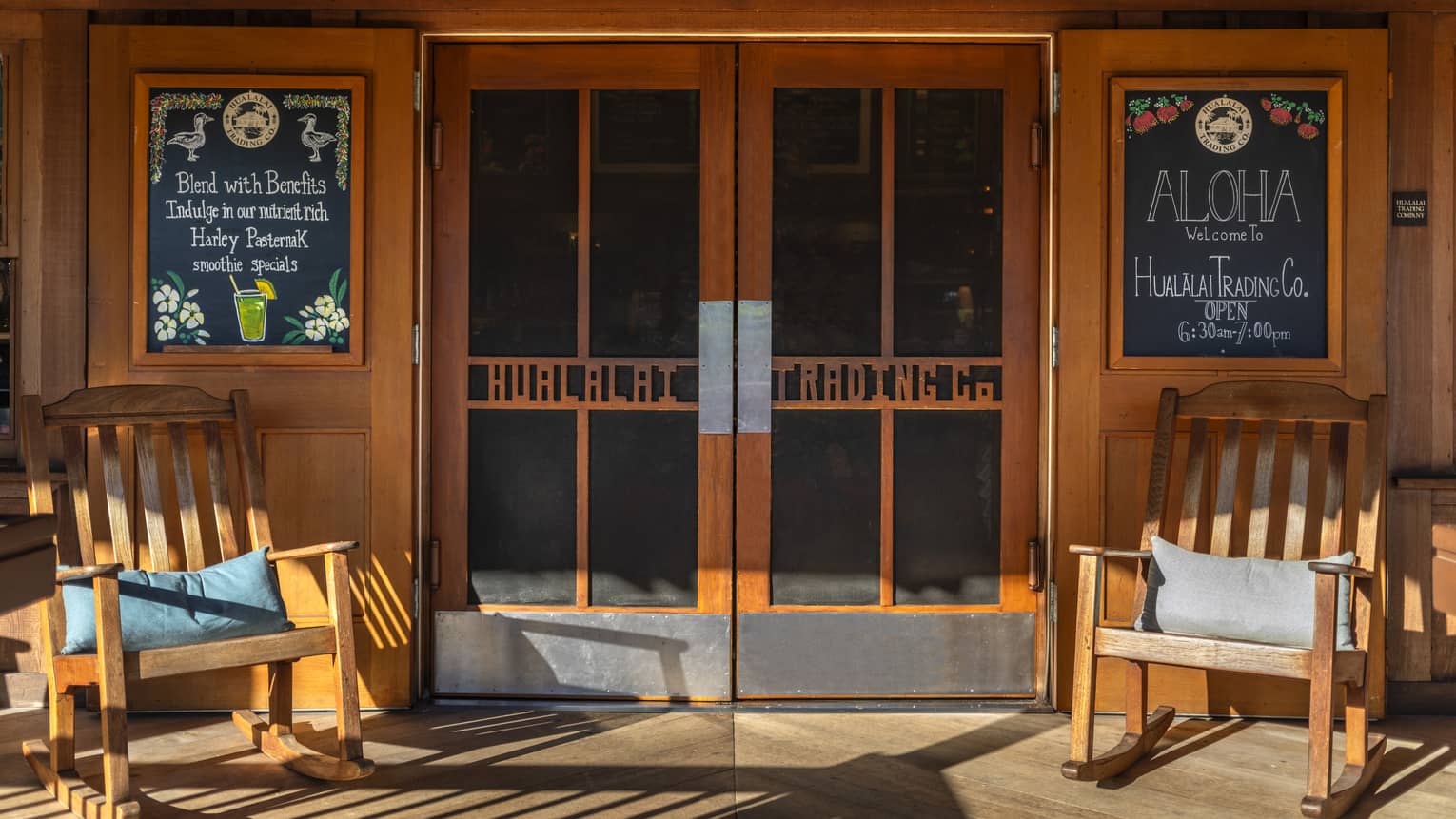 Exterior entrance of a coffee shop and cafe, featuring two wooden rocking chairs and chalk boards reading "Aloha" and showing the day's specials