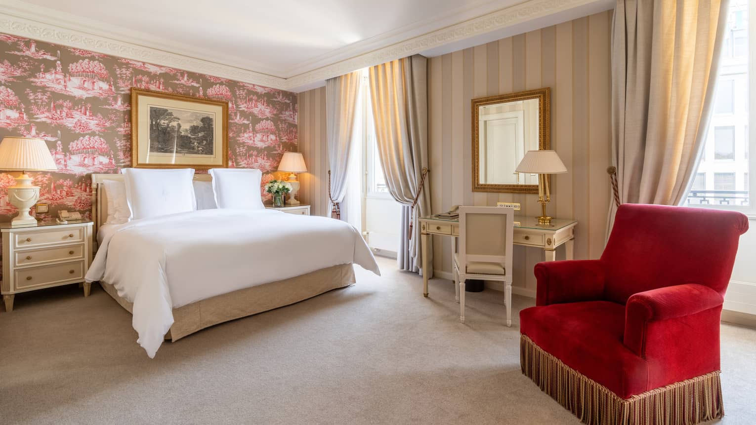 Bedroom with white bed, flanked by nightstands against toile-papered wall with painting, desk and red club chair