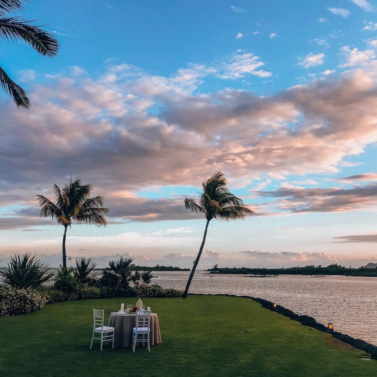 A private dining table for two at sunset, located oceanside on a green patch of grass, surrounded by palm trees and greenery