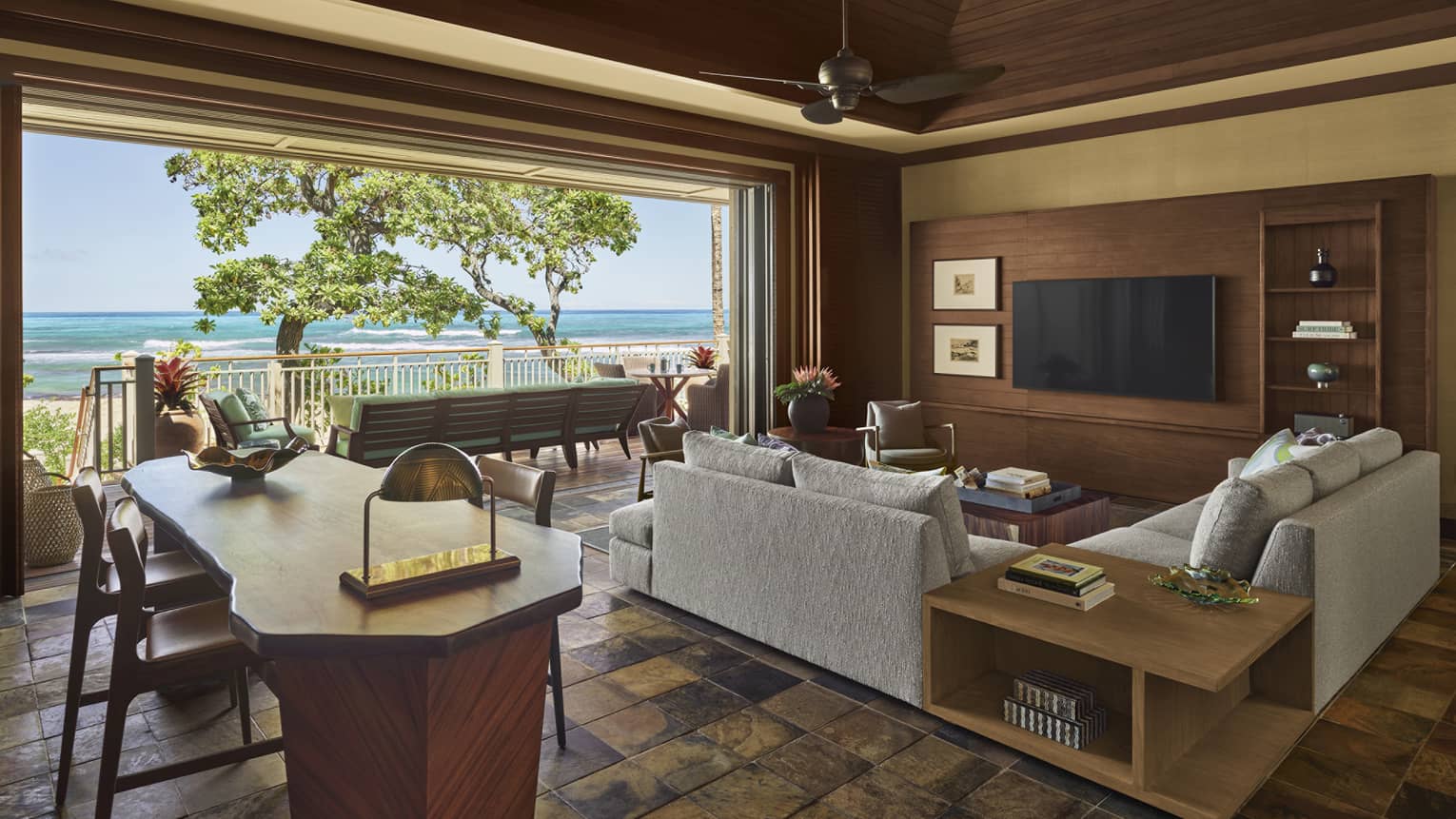 Living area with sectional grey sofa, wall TV, natural wood dining table, opening to beach terrace
