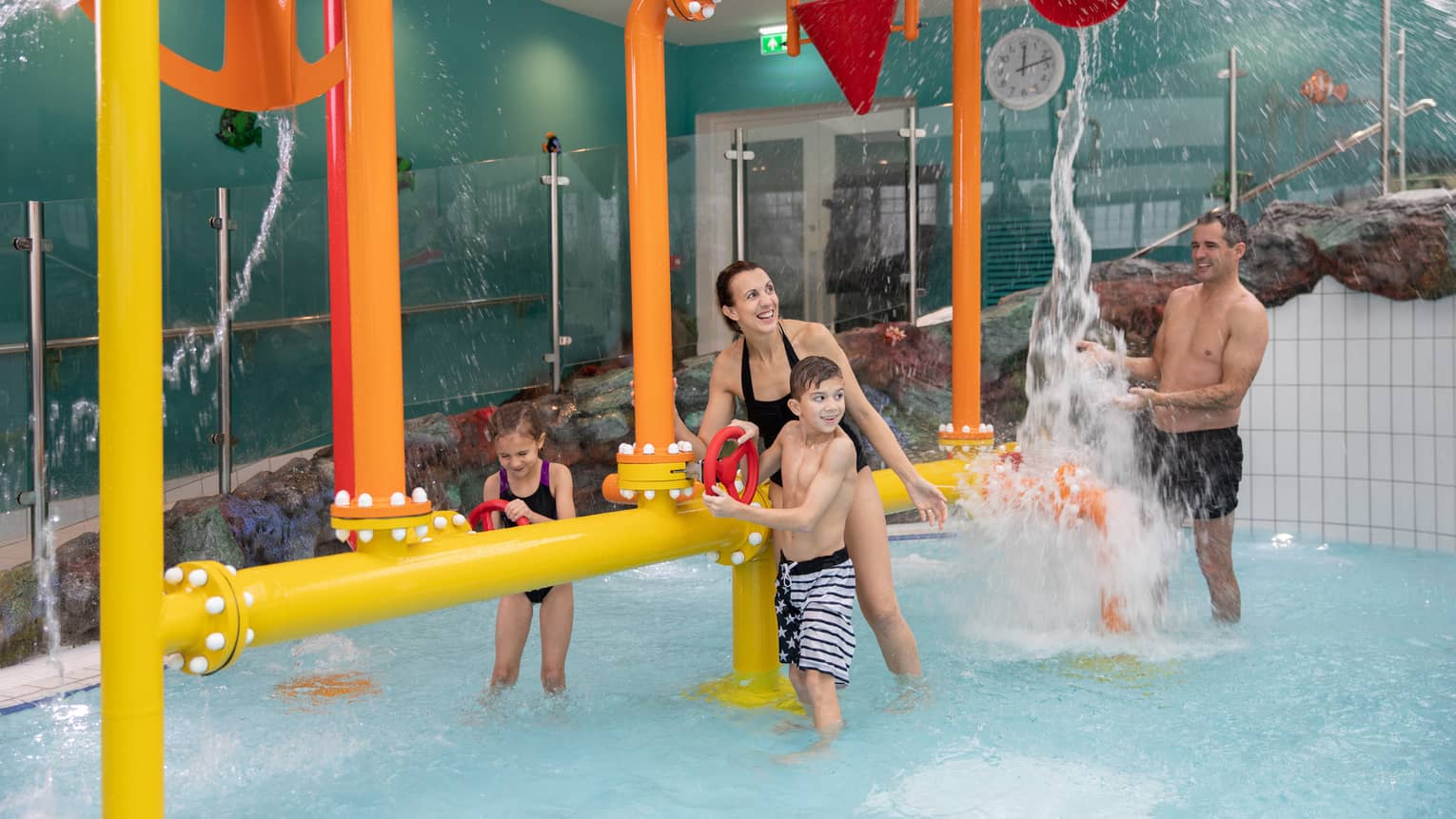 A man, woman and two children play in an indoor family pool with fun water pumps and spouts