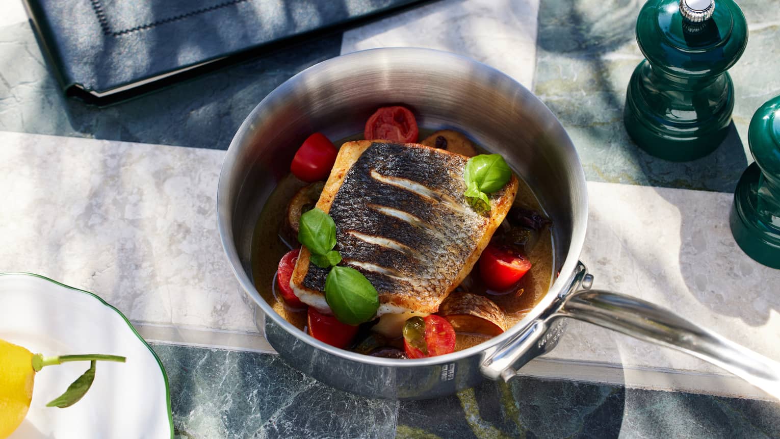 Filet of grilled fish served in a stainless steel pot along with sliced tomatoes and garnished with green leaves