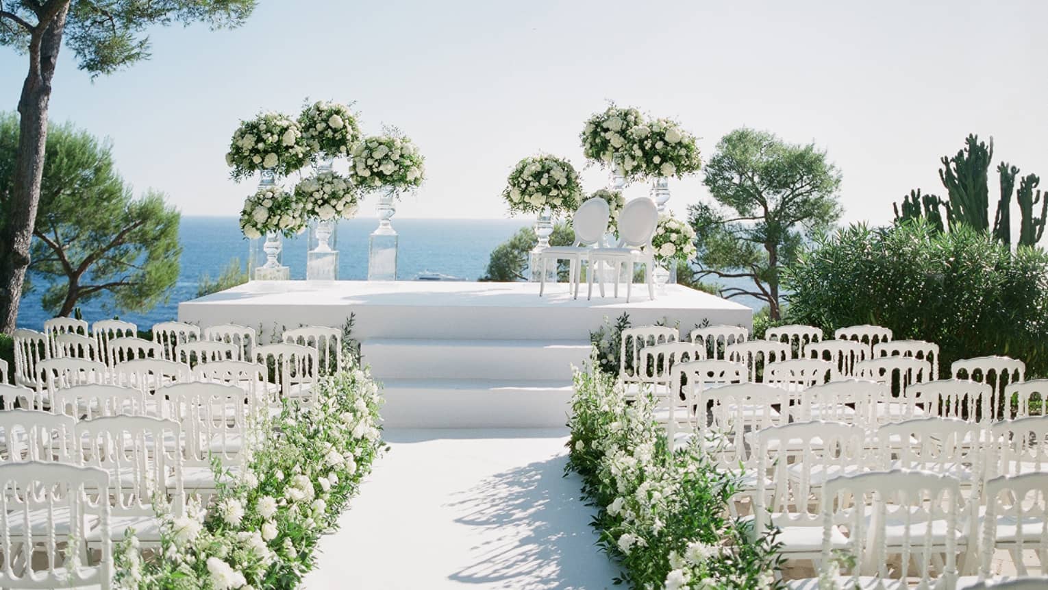 Outdoor wedding setup with white chairs and flower-lined aisle leading up to white altar overlooking Mediterranean Sea