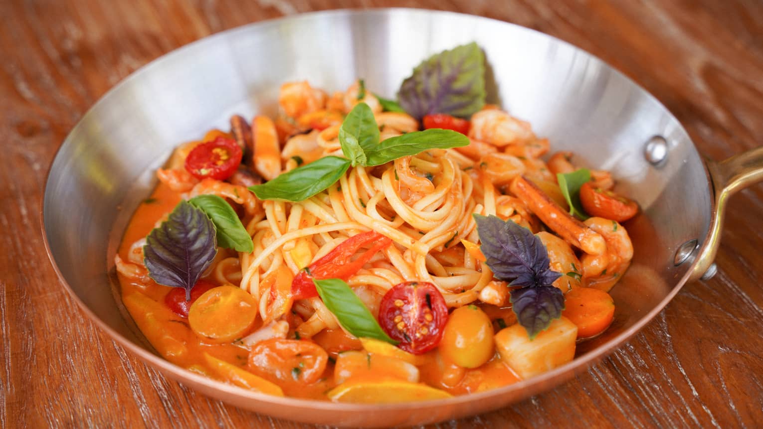 A metal skillet contains long noodles topped with colorful ingredients and garnishes.