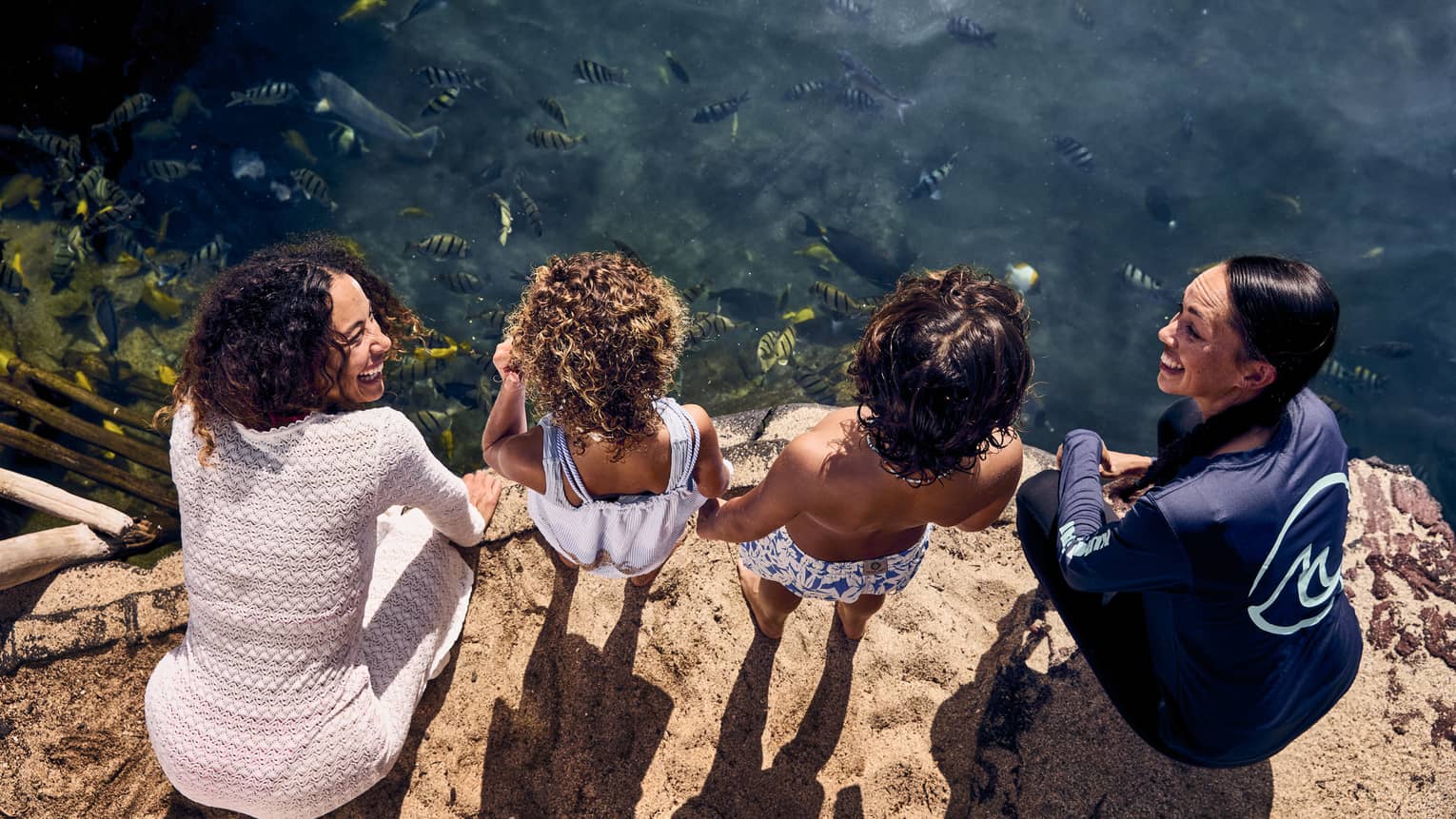 Two women and two young children stand on the edge of a pond with fish in it