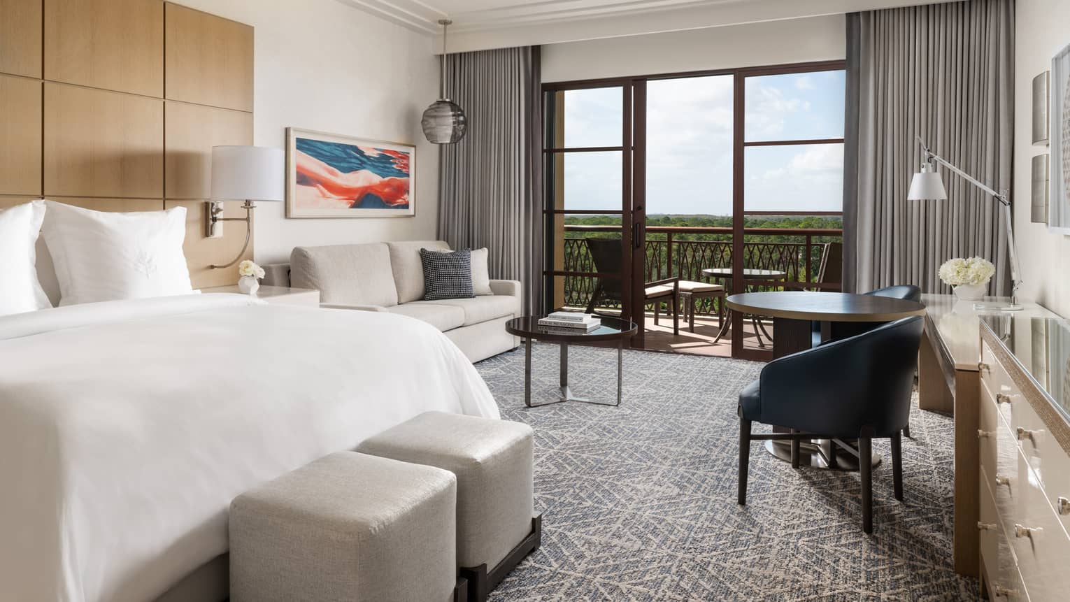 Hotel room with white bed, beige sofa, small table and two black chairs, walk-out balcony