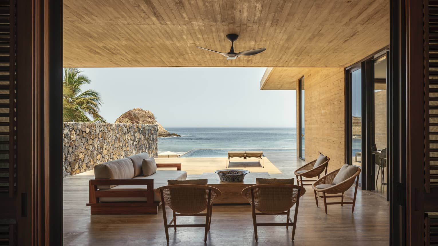 An open-air luxury villa overlooking the ocean, featuring a shaded seating area with modern wooden furniture and a private infinity pool extending towards the beach