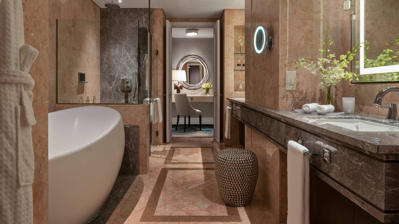 Bathroom with marble walls, featuring a freestanding white bathtub, glass-enclosed shower, double vanity and seating area with a circular mirror.