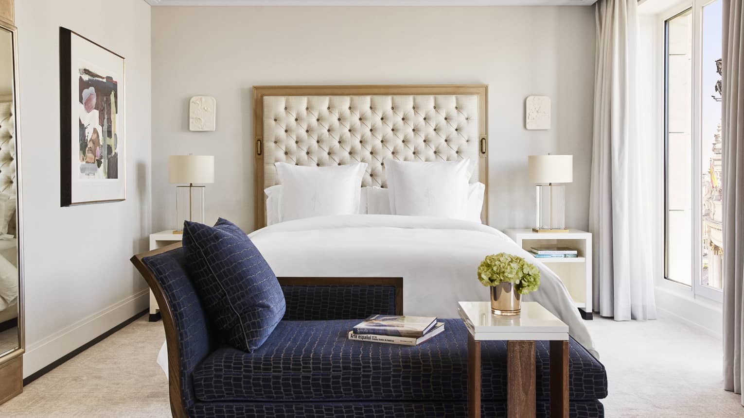 Luxury hotel bedroom with king bed, tufted headboard, blue chaise lounge with books, bedside tables with lamps, wall art and large windows