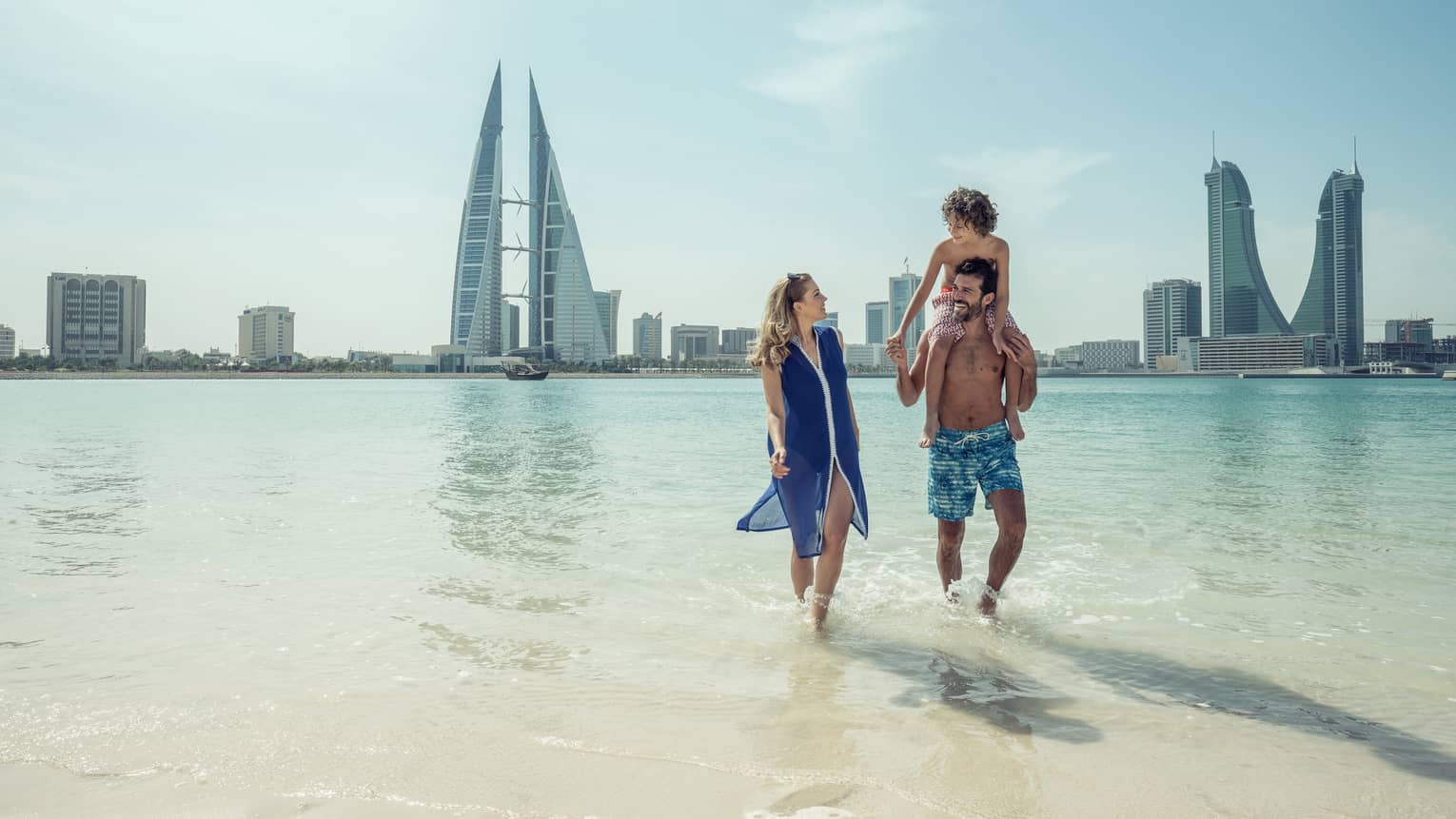A man with a teenager on his shoulders walks with a woman through shallow ocean water with the city of Bahrain Bay in the background