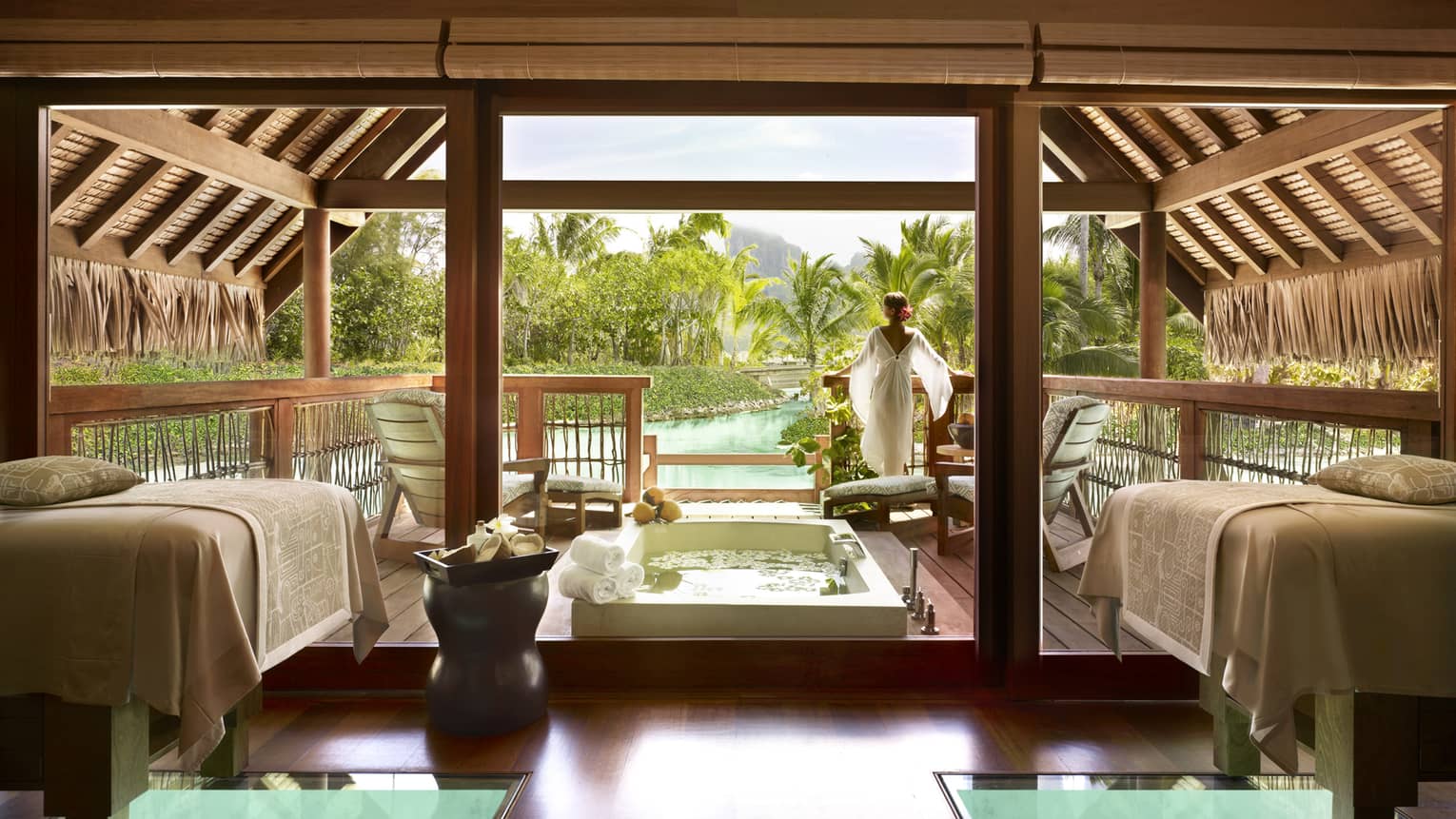 Woman wearing flowing white robe stands at edge of spa suite near plunge bath, couples massage beds
