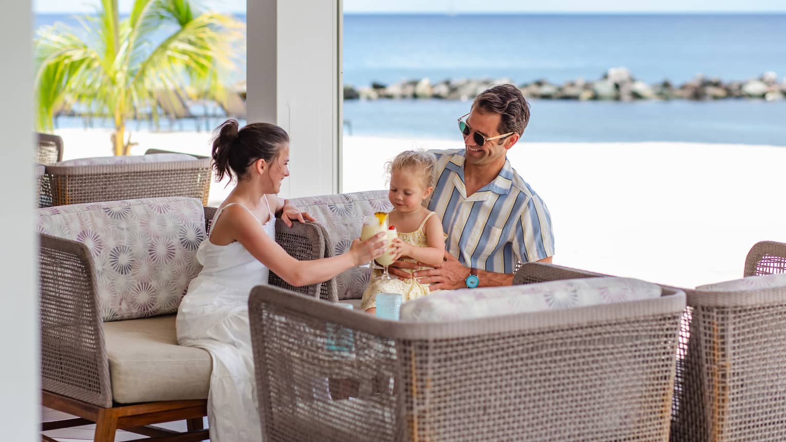 An adult wearing a blue-and-white striped button down short-sleeve shirt holds a young child on his lap, while a young teenager in a white dress sits next to them playing with the baby with the beach in the background
