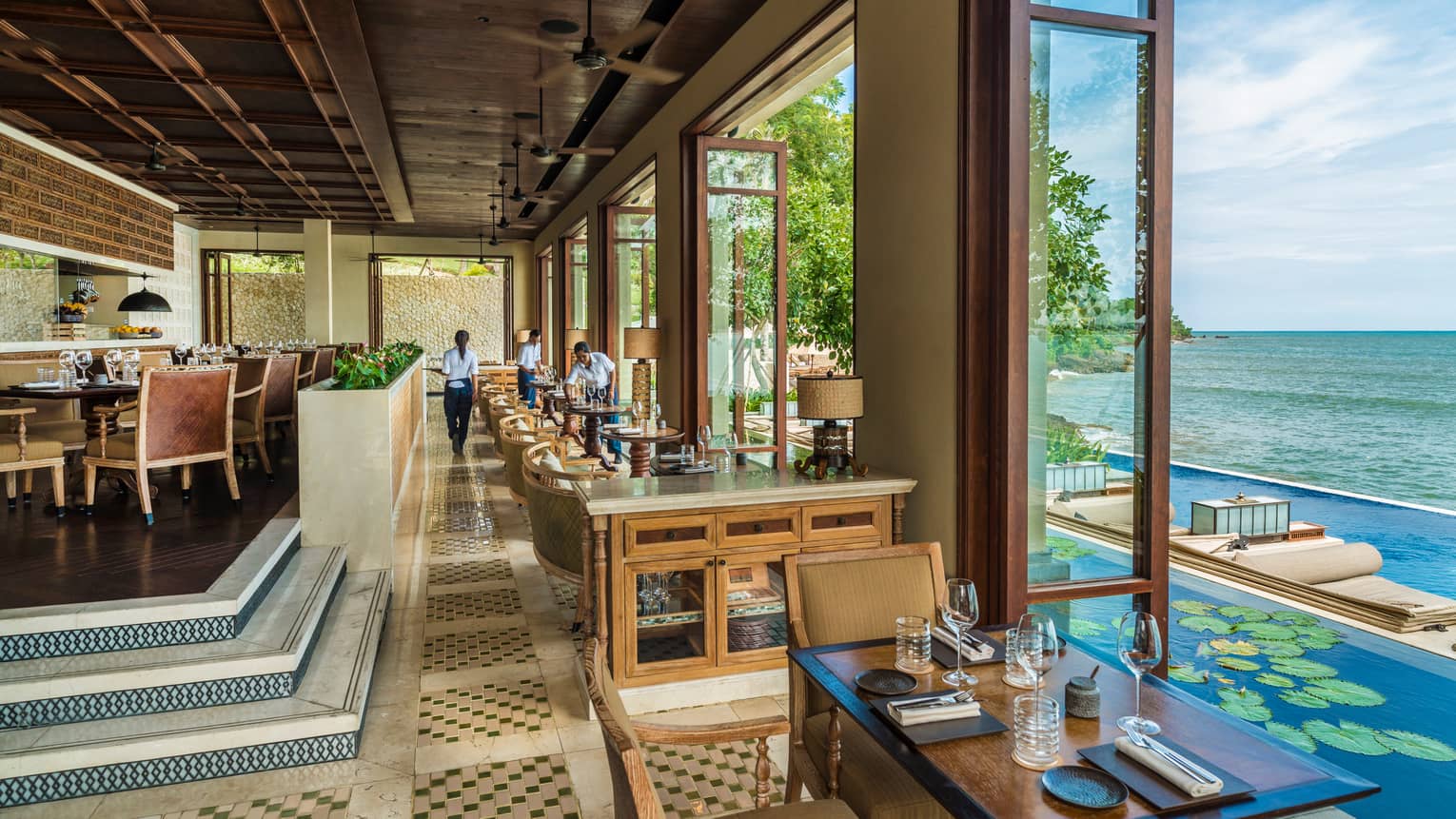 Airy dining room with tables, stairs down to cocktail tables by open floor-to-ceiling windows looking at lotus pond, pool, ocean