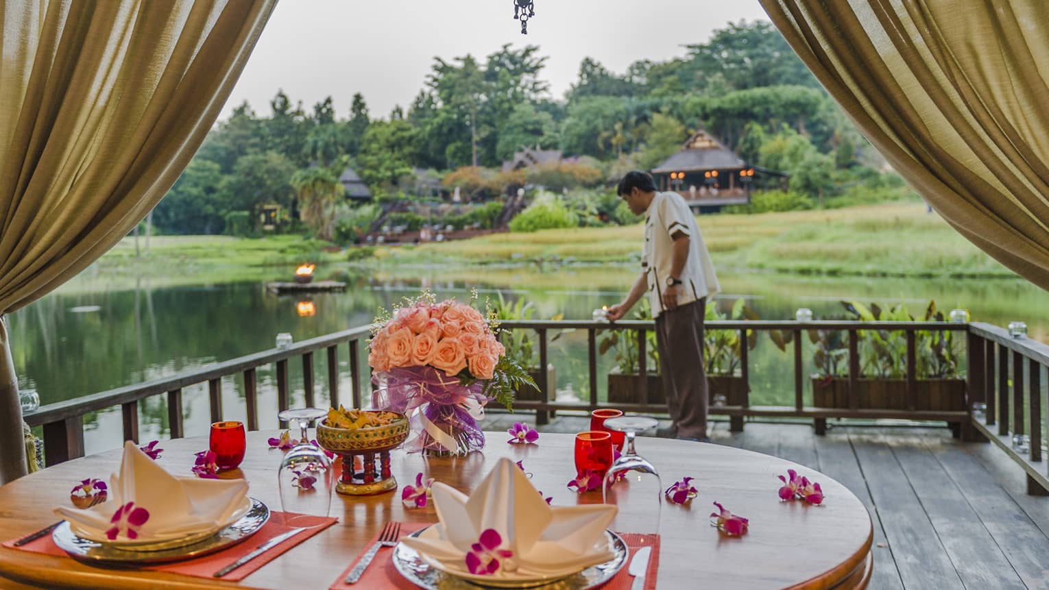 Private dining table with pink flowers, napkins on pavilion, hotel staff lights candle near water 