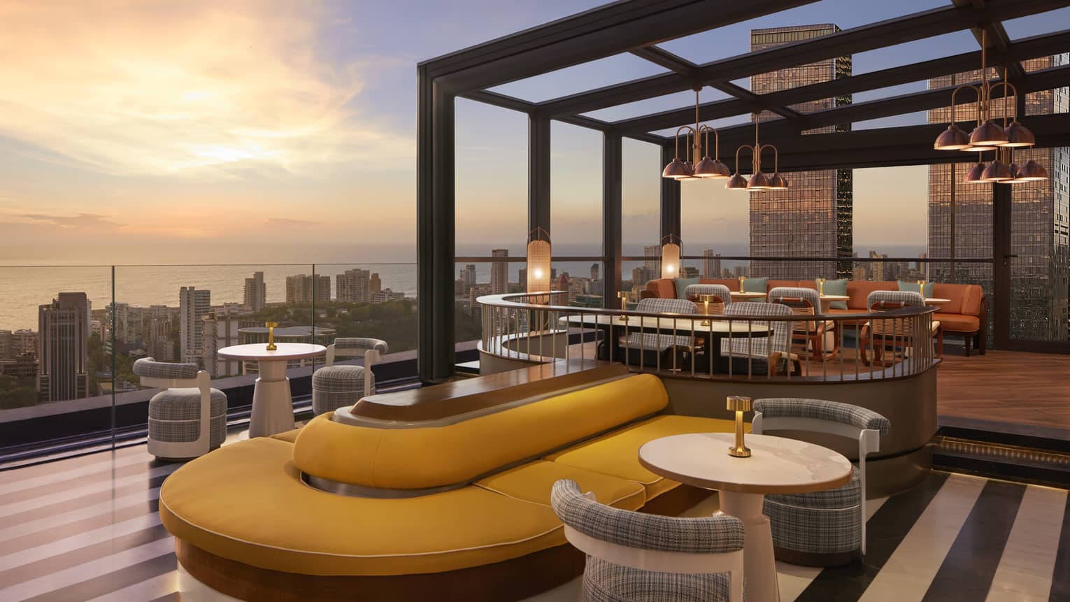 A modern rooftop patio with a mix of booth bank seating and tables for two, panoramic cityscape view and dusky ocean beyond.