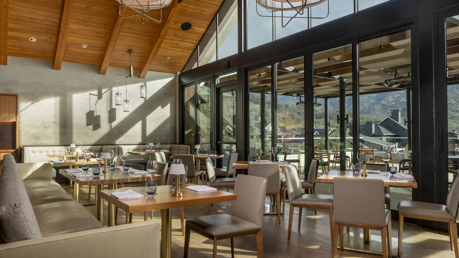 Modern restaurant interior with high wooden ceilings, large windows, and natural light. Tables are set, and an outdoor terrace offers views of surrounding hills.