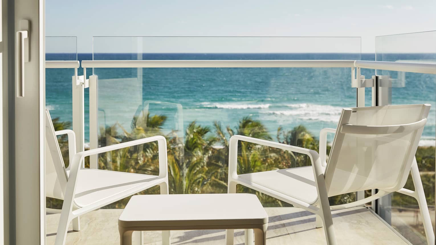 Luxury hotel balcony with two white chairs and a table overlooking the ocean and palm trees under a clear blue sky