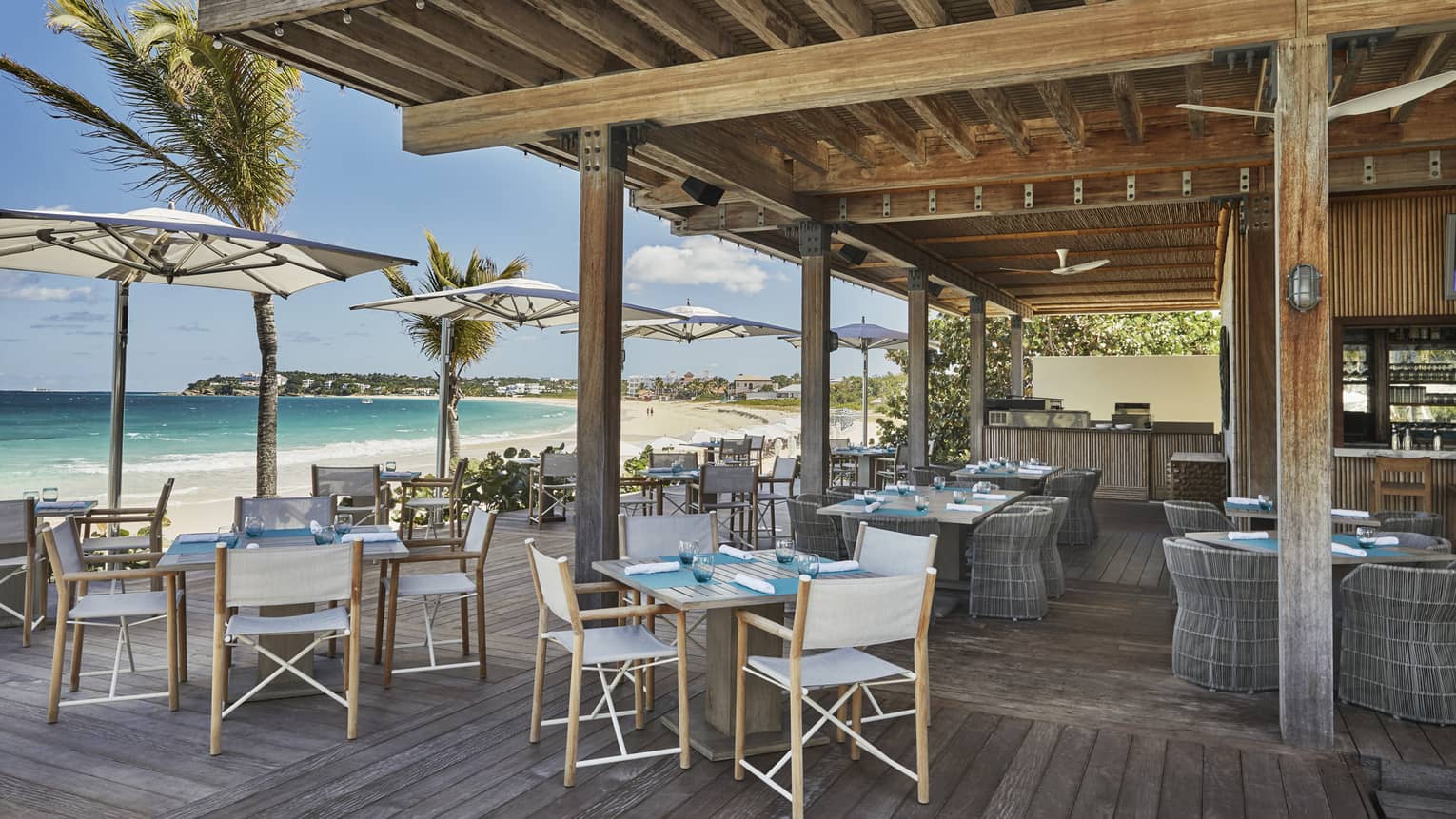 Outdoor beachfront restaurant with wooden floors, umbrella-shaded tables and ocean views.