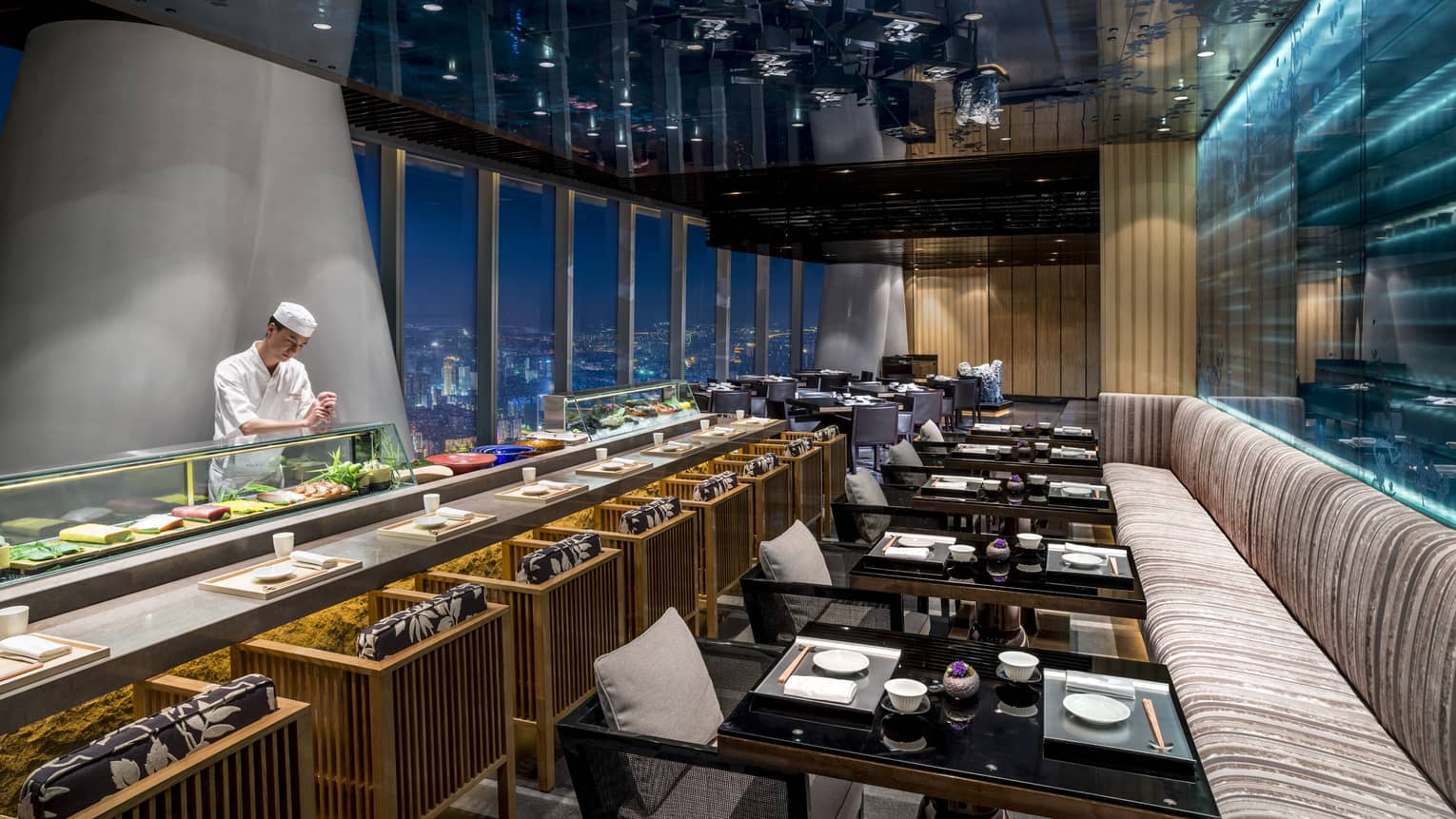 Kumoi Japanese restaurant chef stands behind long sushi bar in dining room overlooking city lights