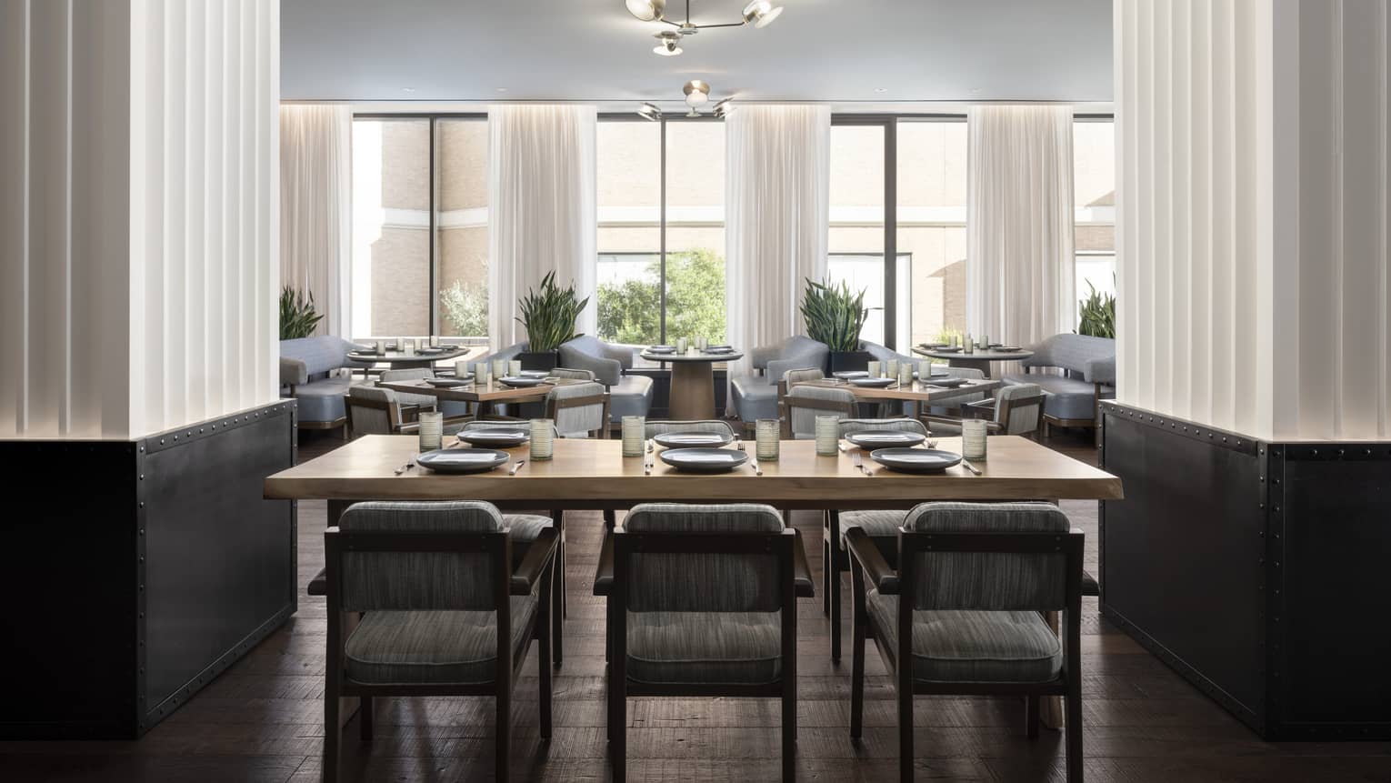 A dining area with a wood rectangular table and grey cloth chairs, large windows can be seen in the background.