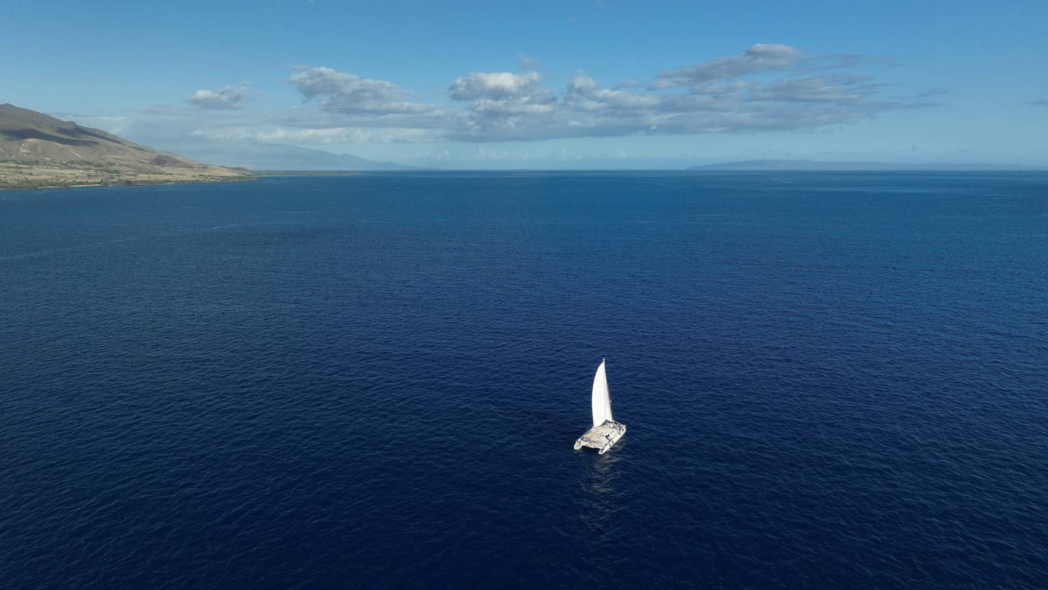 Aerial view of a catamaran as it sails through the vast, calm open waters toward the faded silhouette of distant mountains.