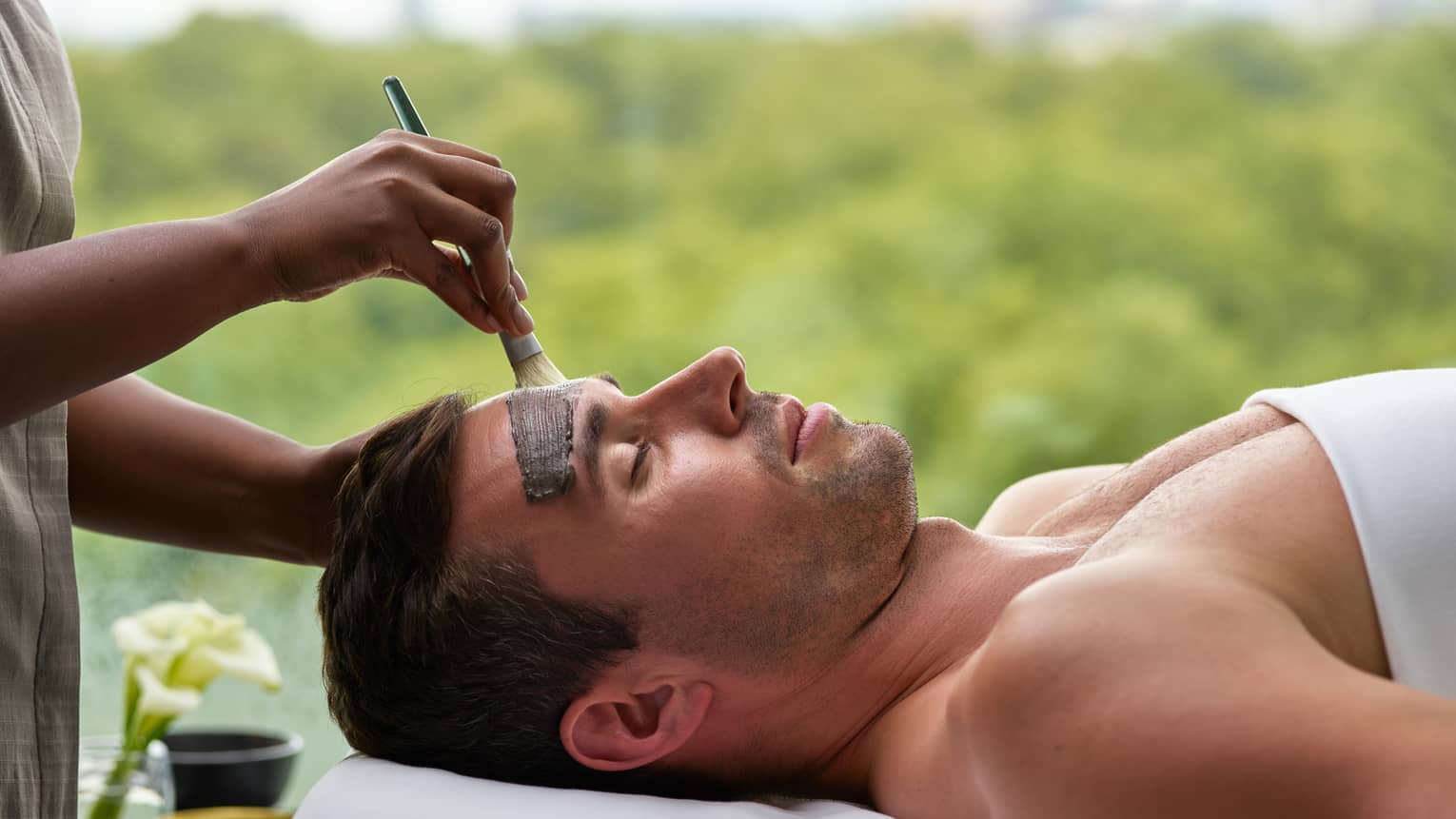 Man lying under white sheet on spa table as staff paints clay mask on his forehead