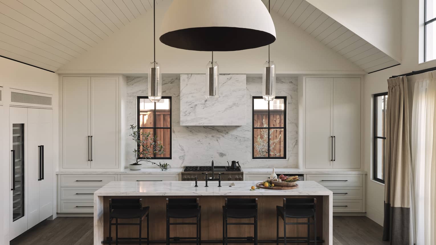 Luxury resort kitchen with vaulted high ceiling, marble island, black barstools, large white cabinets, stove, pendant lights and windows