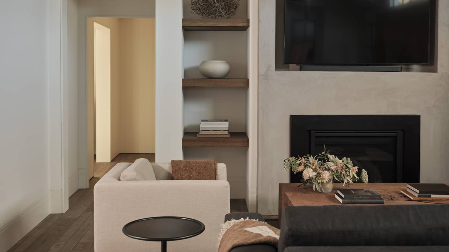 Living room with gray sofa, beige armchair, side table, wooden coffee table with floral arrangement, built-in shelves with decor, wall-mounted TV and fireplace