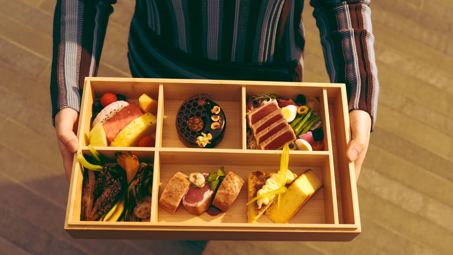 A person holds a wooden bento box?style tray with fine food items in it