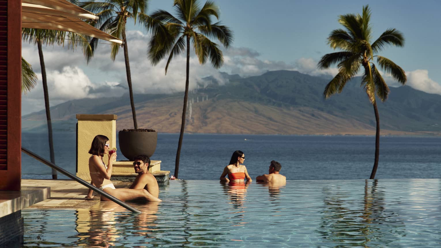 Four people relax in an infinity pool overlooking the ocean and palm trees with a mountain in the background