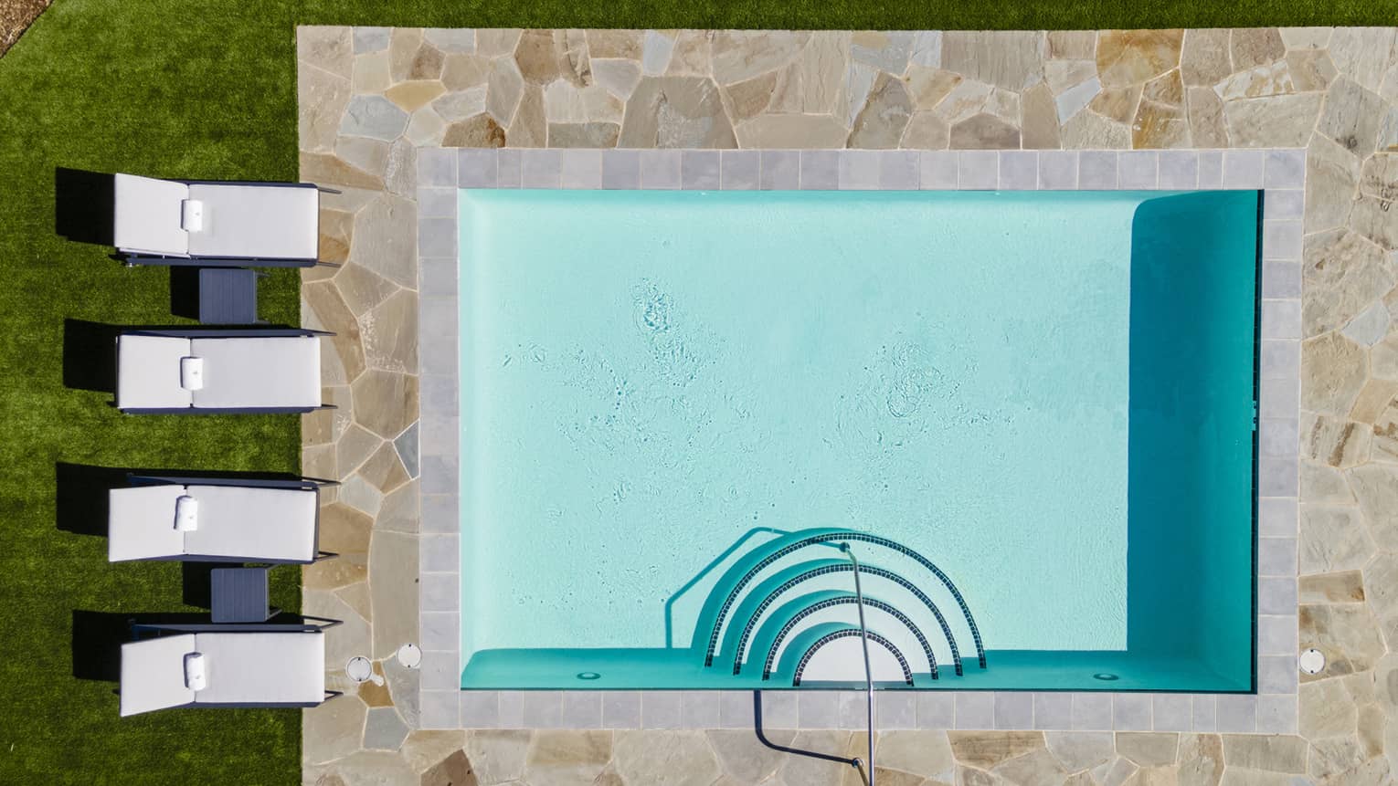 Aerial view of a small rectangular swimming pool with stone tile surround, adjacent to sun loungers on green grass