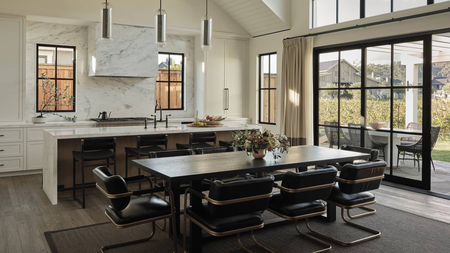 Open kitchen and dining area with marble island, dark wood table, black leather chairs, pendant lights, windows and sliding doors leading to outdoor patio