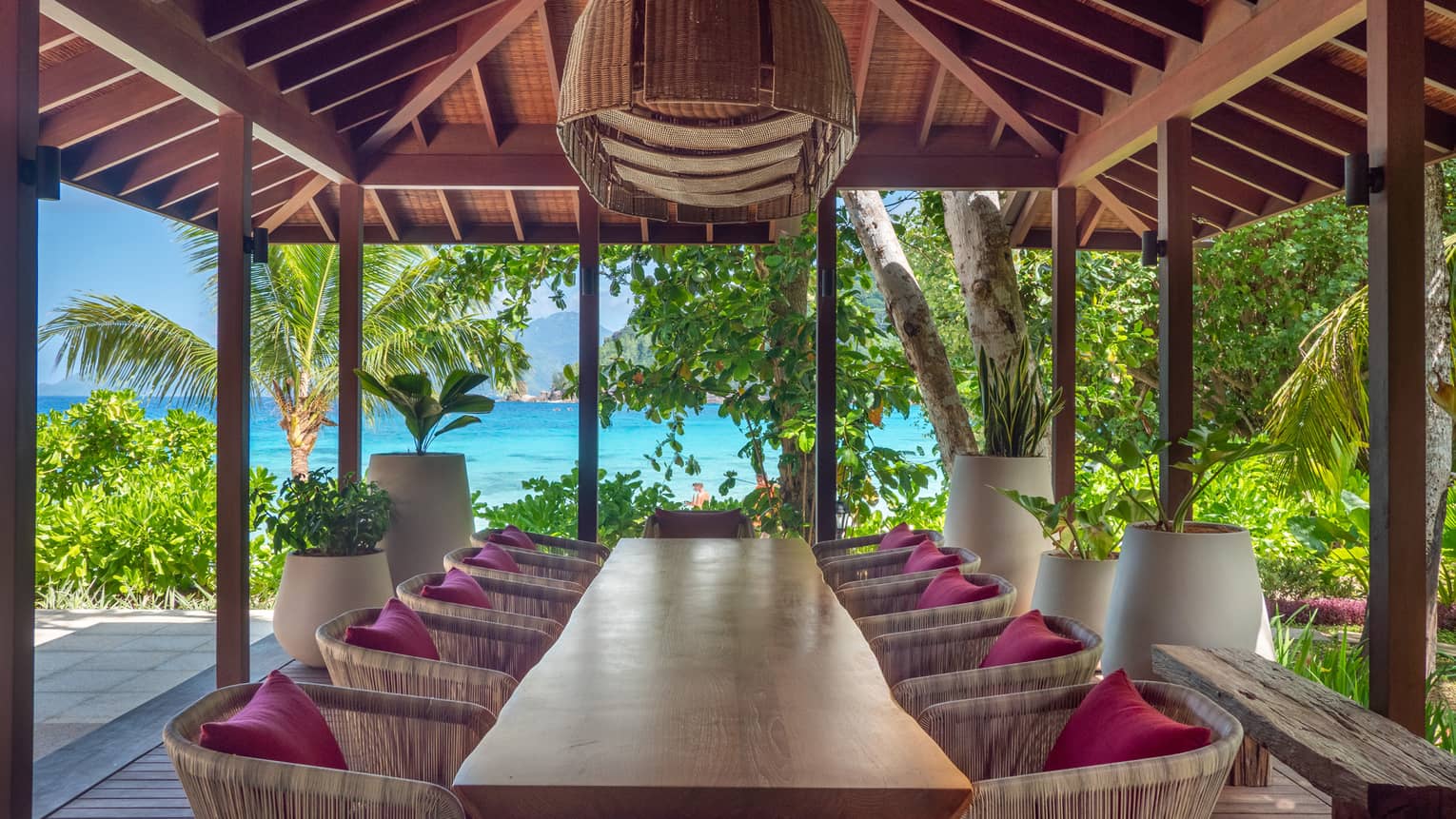 Natural wood dining table with 10 chairs underneath an outdoor pavilion with sea view