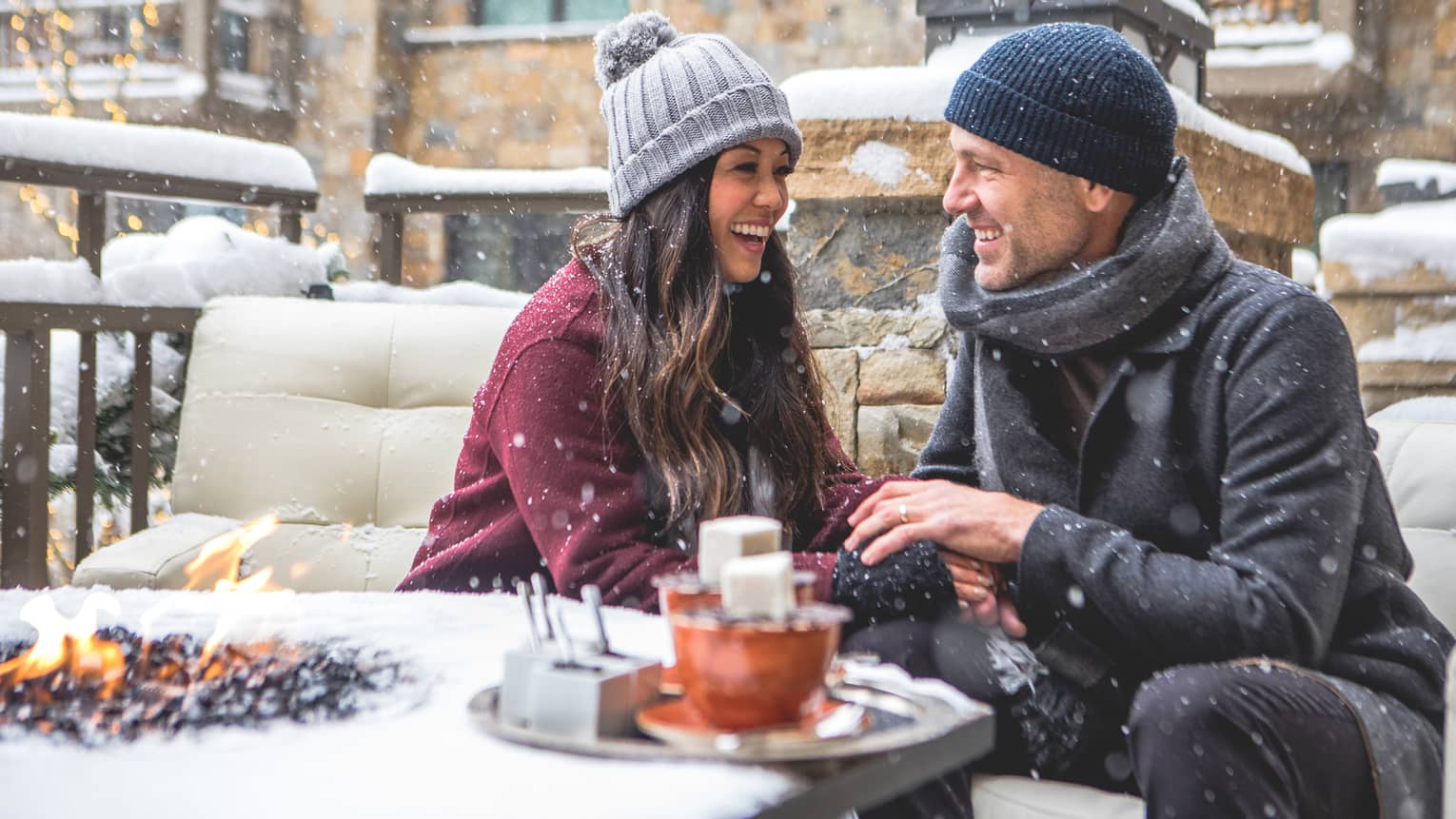 Smiling couple sitting by a firepit in a snowy outdoor setting, bundled in winter coats and hats. Snowflakes fall around them as they enjoy a cosy moment. 