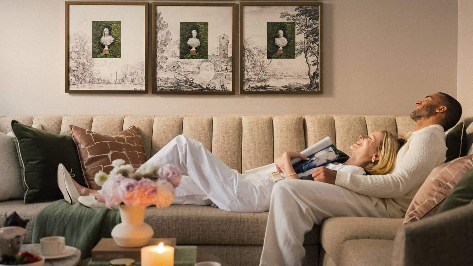 Two guests laying on the couch in a guest room with picture frames on the wall and a bouquet of flowers on the coffee table