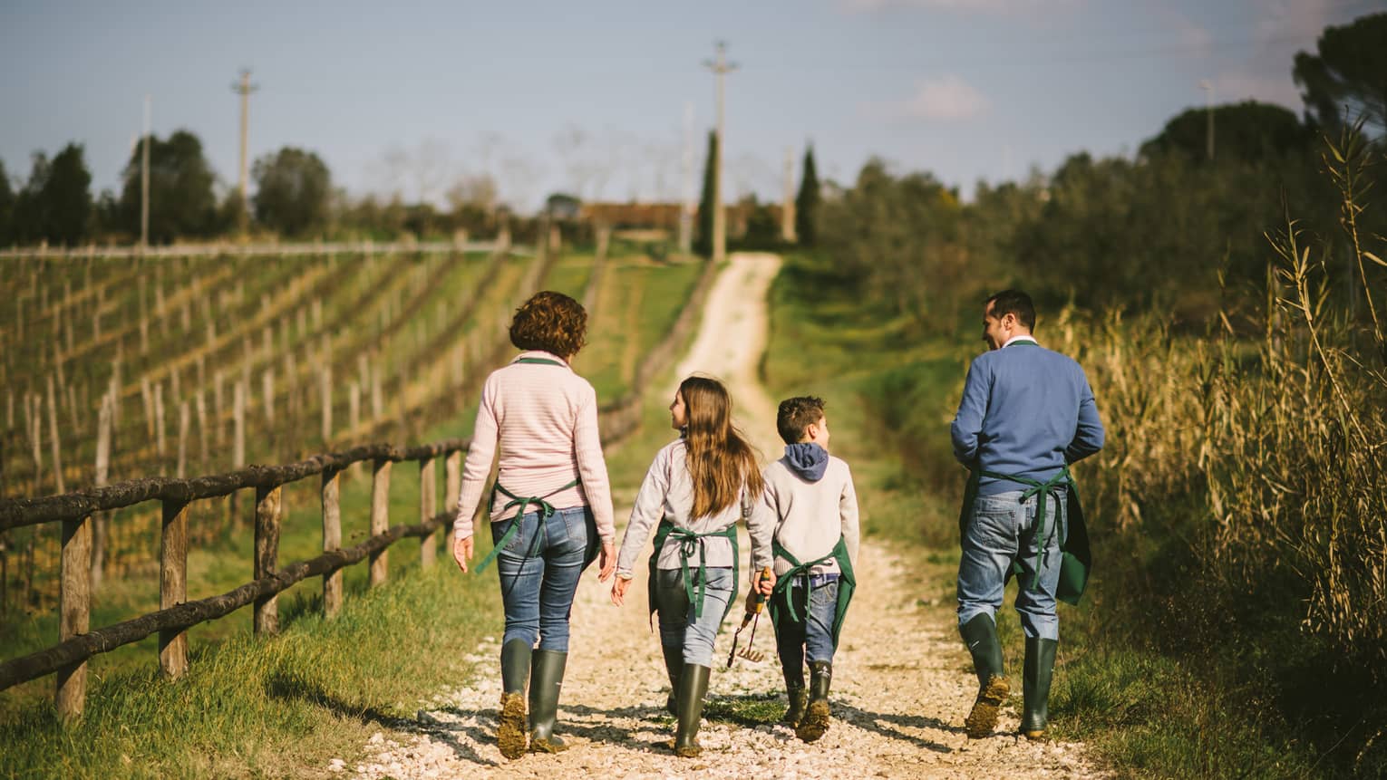 A mother, father, daughter, and son walking along a wood fence on a dirt path with wheat plants next to them.