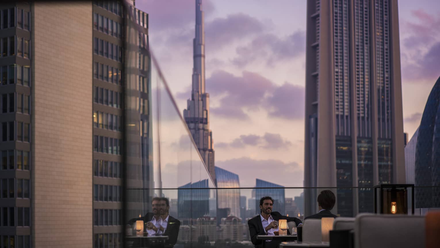 Men in suits on rooftop patio in front of glass balcony wall overlooking city at sunset