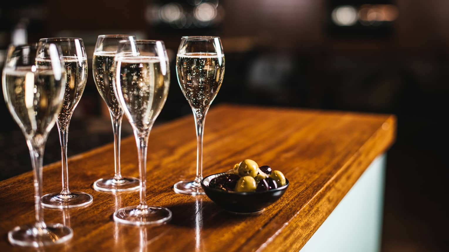 Five glasses of sparkling wine with bowl of black and green olives, atop a wooden bar top
