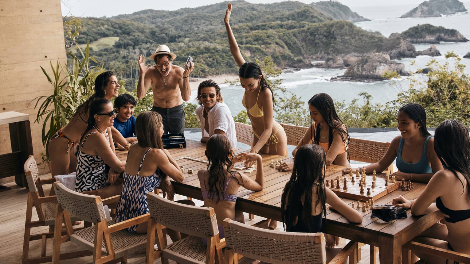 A group of family and friends enjoying time together at a wooden table, playing chess and laughing, with a scenic view of the ocean and lush green hills in the background