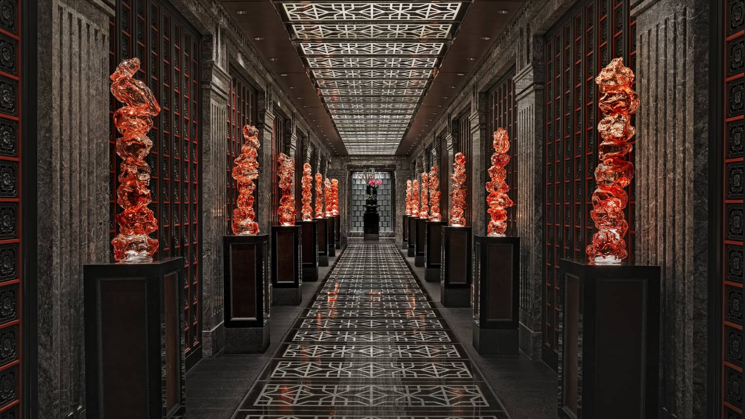 View down a long black-tiled hall lined with orange glass sculptures on pedestals, the geometric ceiling mirroring the floor.