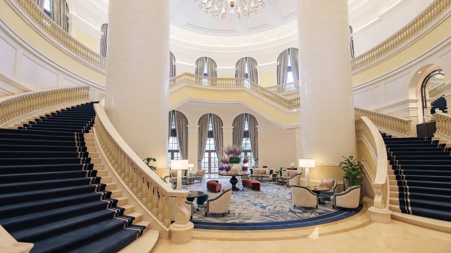 Lobby with winding carpeted staircase on either side of two-storey sitting area, ornate chandelier above and marble flooring