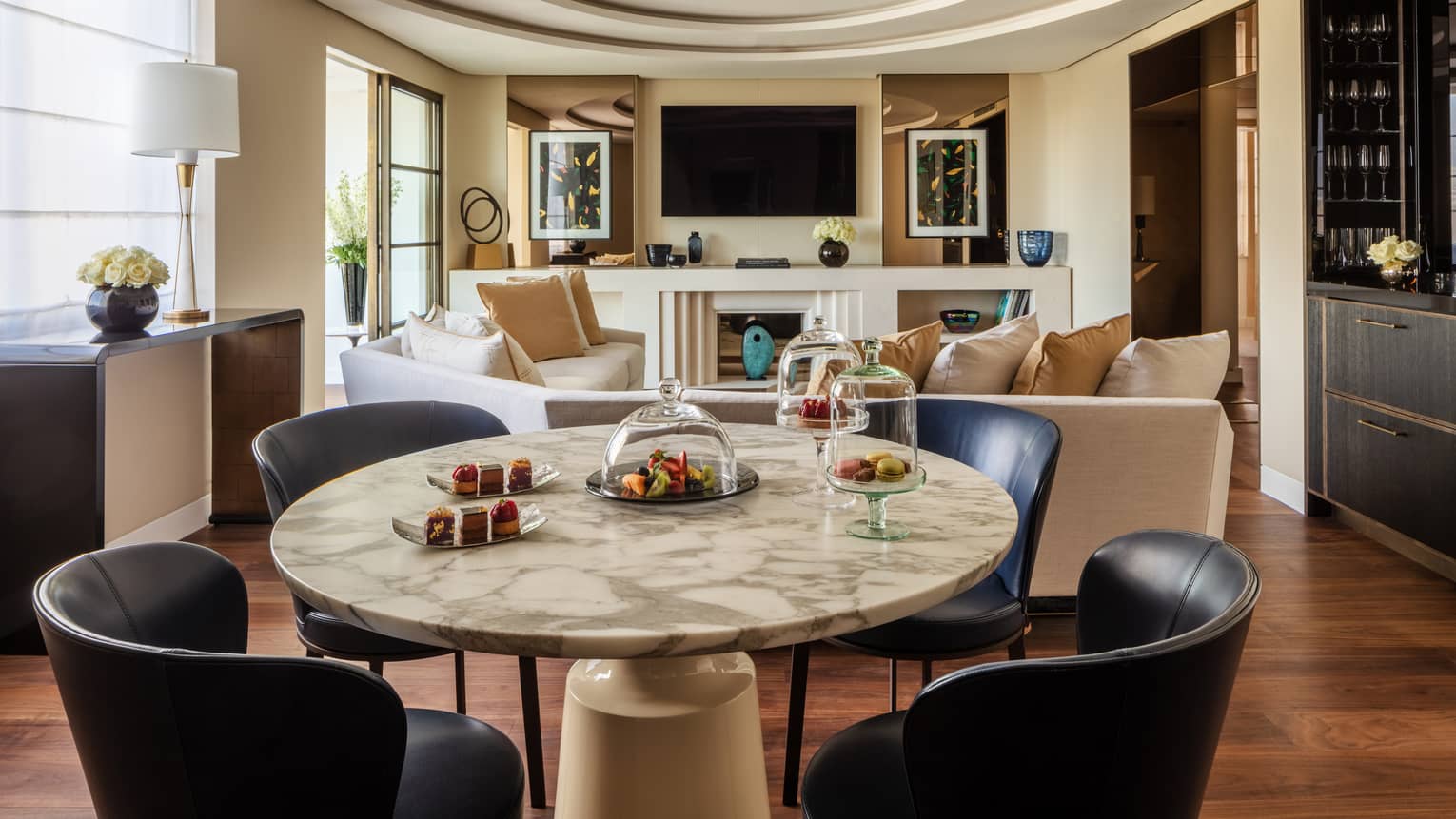 A suite with a round stone table with black chairs, a beige sofa, a television and large windows.